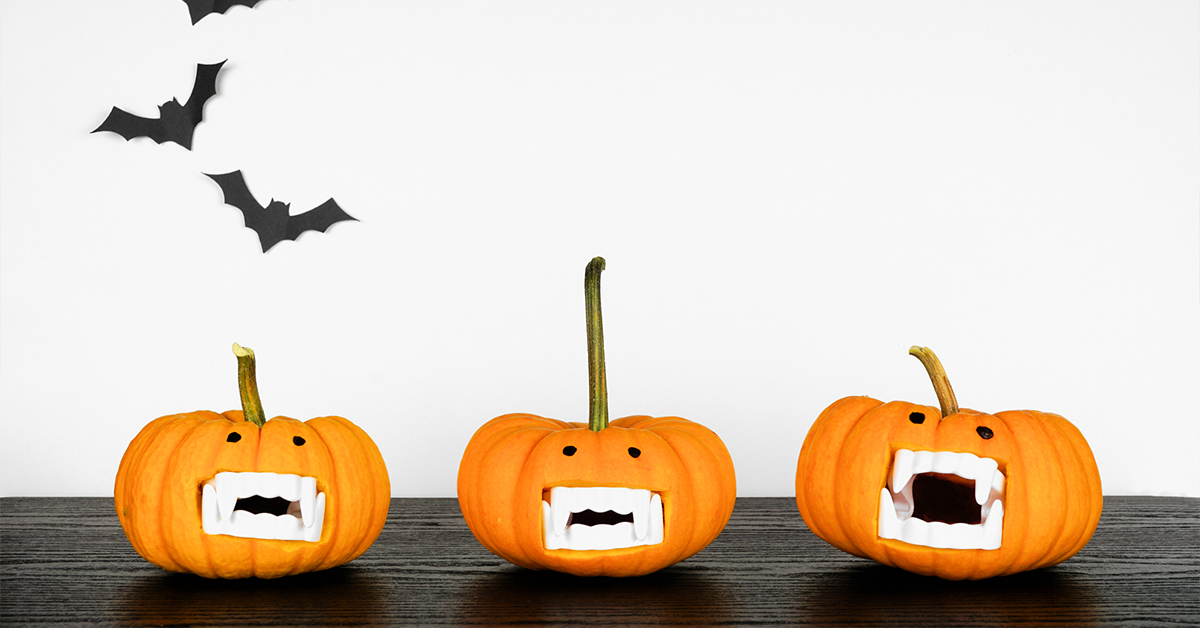 Three orange pumpkins on a table with plastic teeth as the mouth and bats on the wall behind