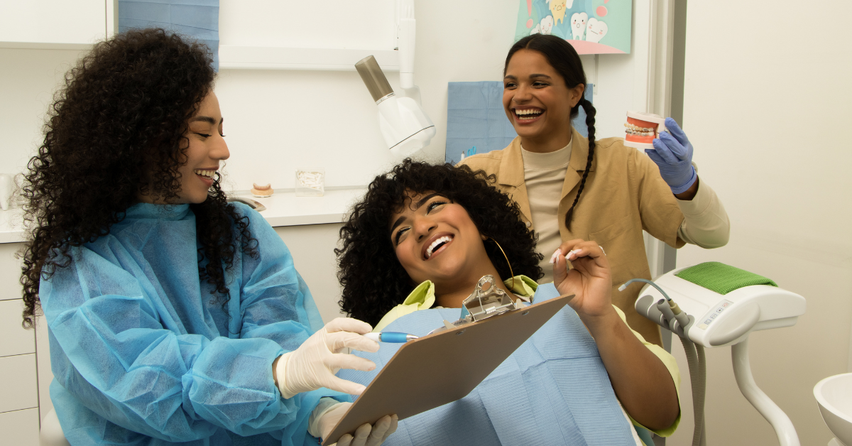 Three women in a dental office smiling at one another. One woman is sitting in a dentists chair looking at a clipboard smiling while another woman smiles and points at the clipboard.